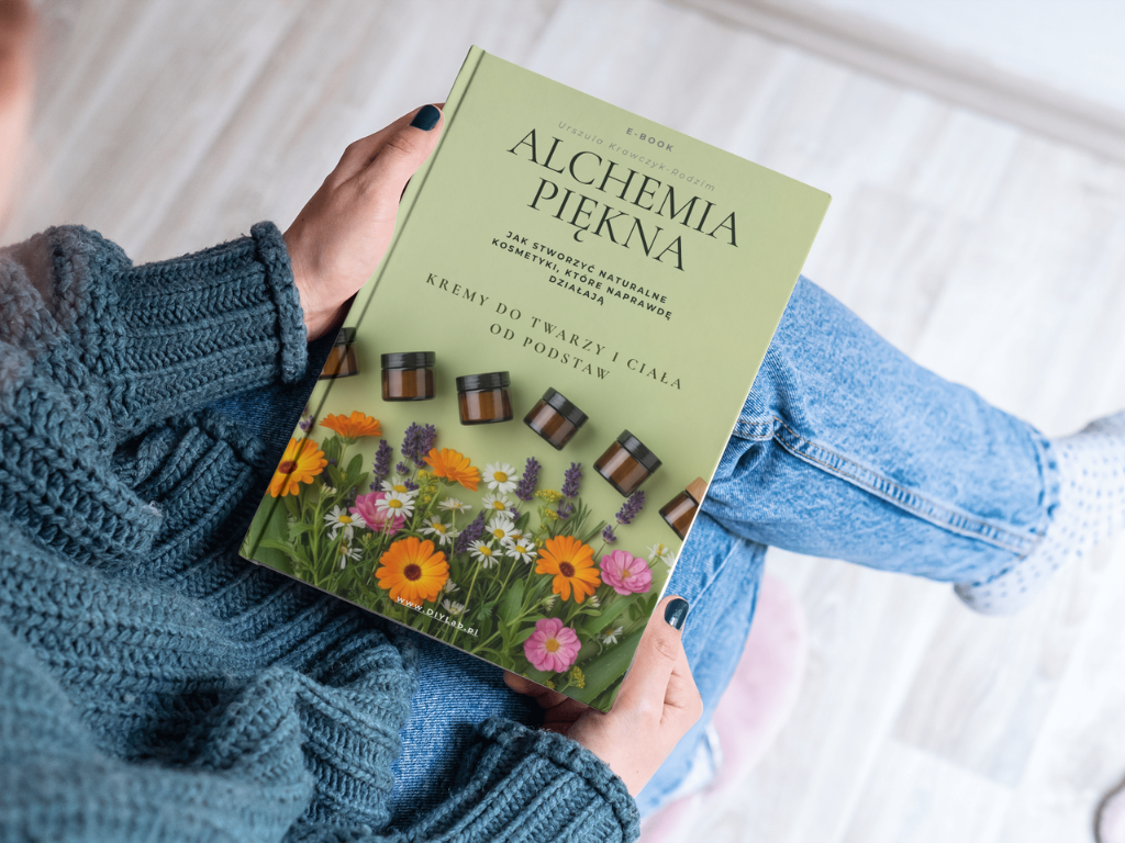kobieta trzyma na kolanach e-book z tytułem "Alchemia Piękna jak zrobić naturalne kosmetyki które działają, kremy do twarzy i cała od podstaw" zielone tło słiczki szklane z ciemnego szkła i kwiaty rożlin leczniczych